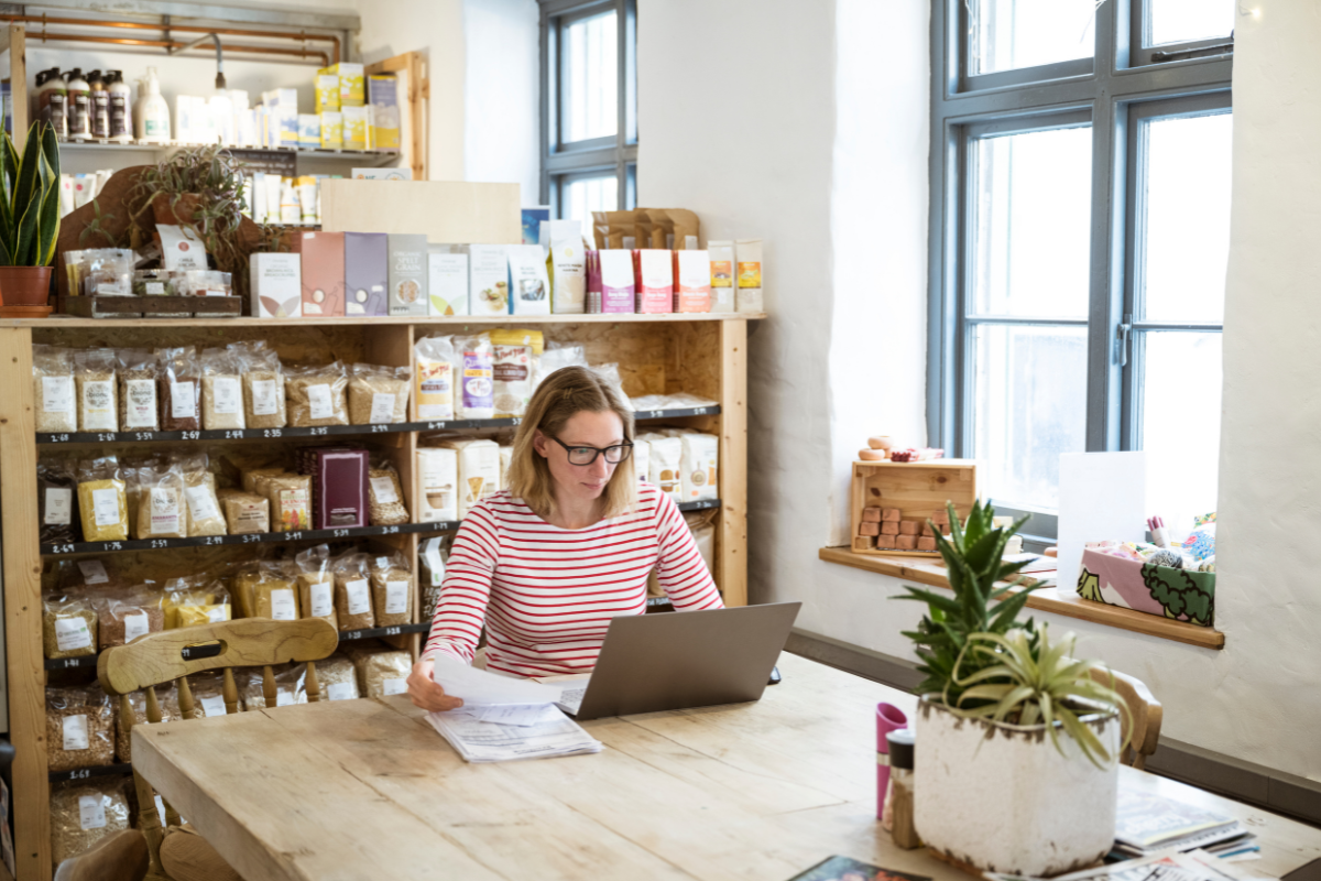 Woman with striped shirt and glasses sits at a wooden table with a laptop reviewing papers in a shop with shelves of groceries behind her