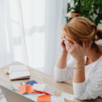 Frustrated woman sitting at a desk with receipts a calculator and a laptop head in hands