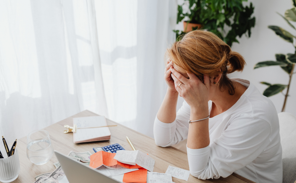 Frustrated woman sitting at a desk with receipts a calculator and a laptop head in hands