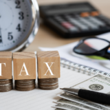 Blocks spelling TAX sit on coin stacks beside a clock calculator glasses and a pen over a notebook with cash nearbyillustrates taxes and accounting